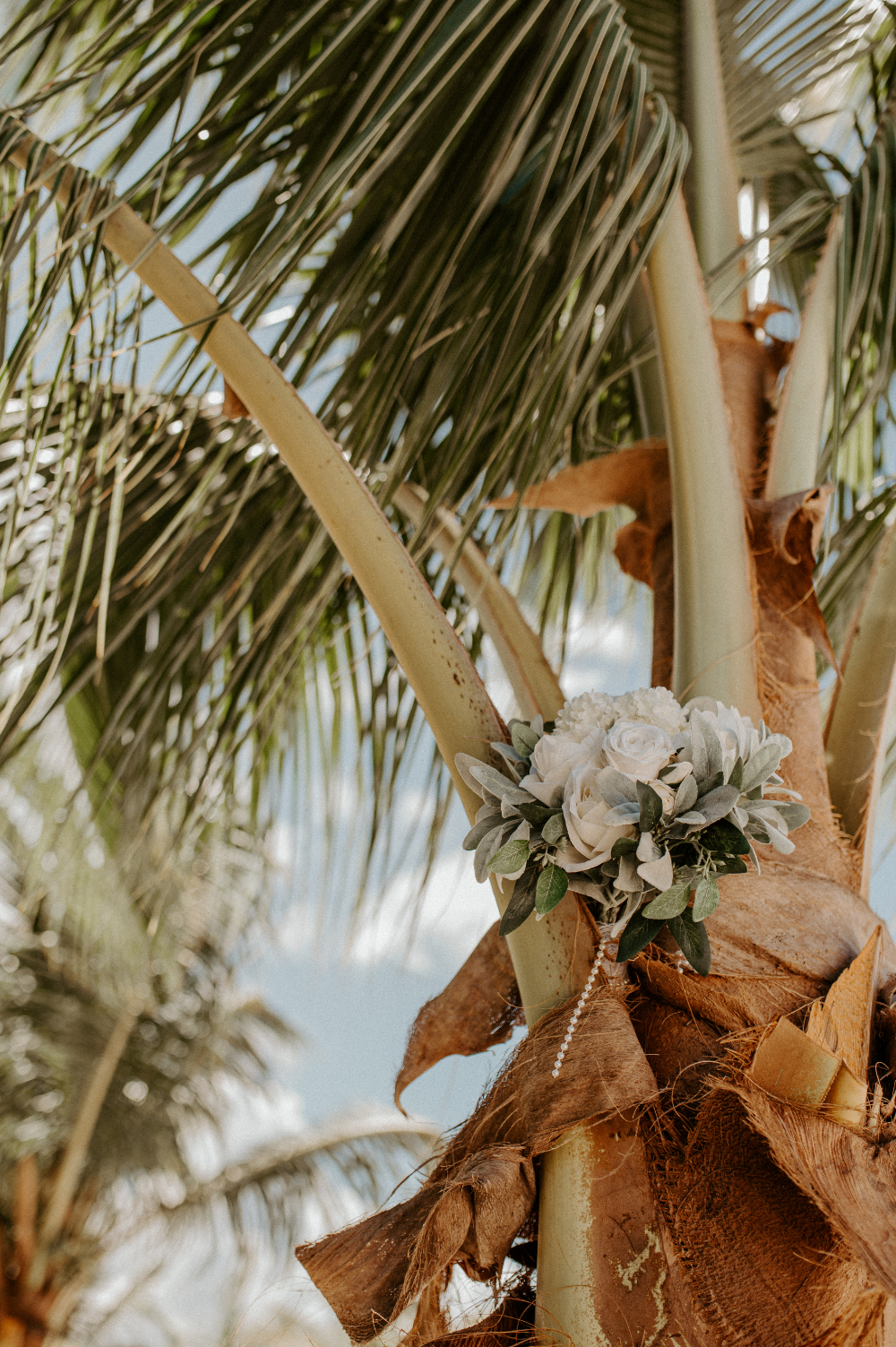 Tropical wedding bouquet tied to palm tree with soft neutral flowers and beachside ceremony decor