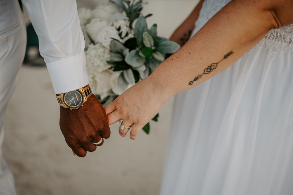 Close-up of couple holding hands with wedding rings and bouquet during intimate beach ceremony