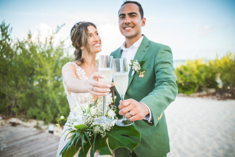 Bride and groom toasting champagne on beach, smiling with bouquet and greenery background