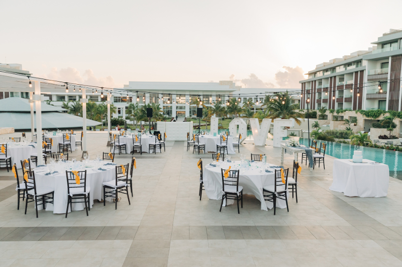 Outdoor destination wedding reception setup with round tables, poolside venue, string lights, and modern resort backdrop