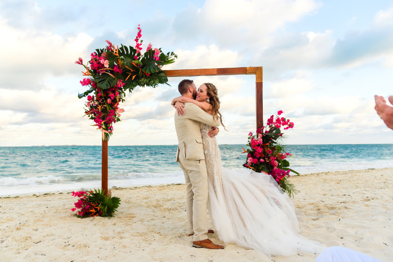 Destination wedding planner helping couple celebrate beachfront wedding ceremony with tropical floral arch at sunset.