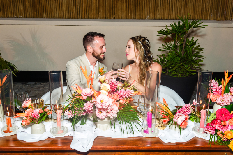 Bride and groom sharing a toast at tropical wedding reception with colorful floral centerpieces and candles.